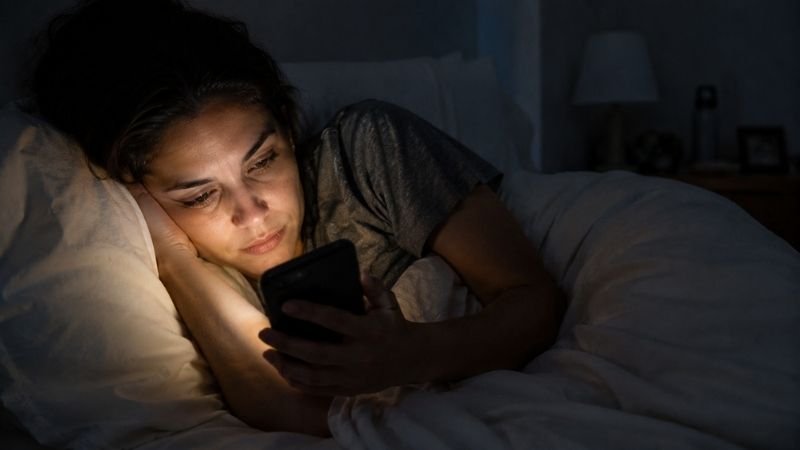 Tired woman scrolling phone late at night, disrupting sleep and weight loss progress